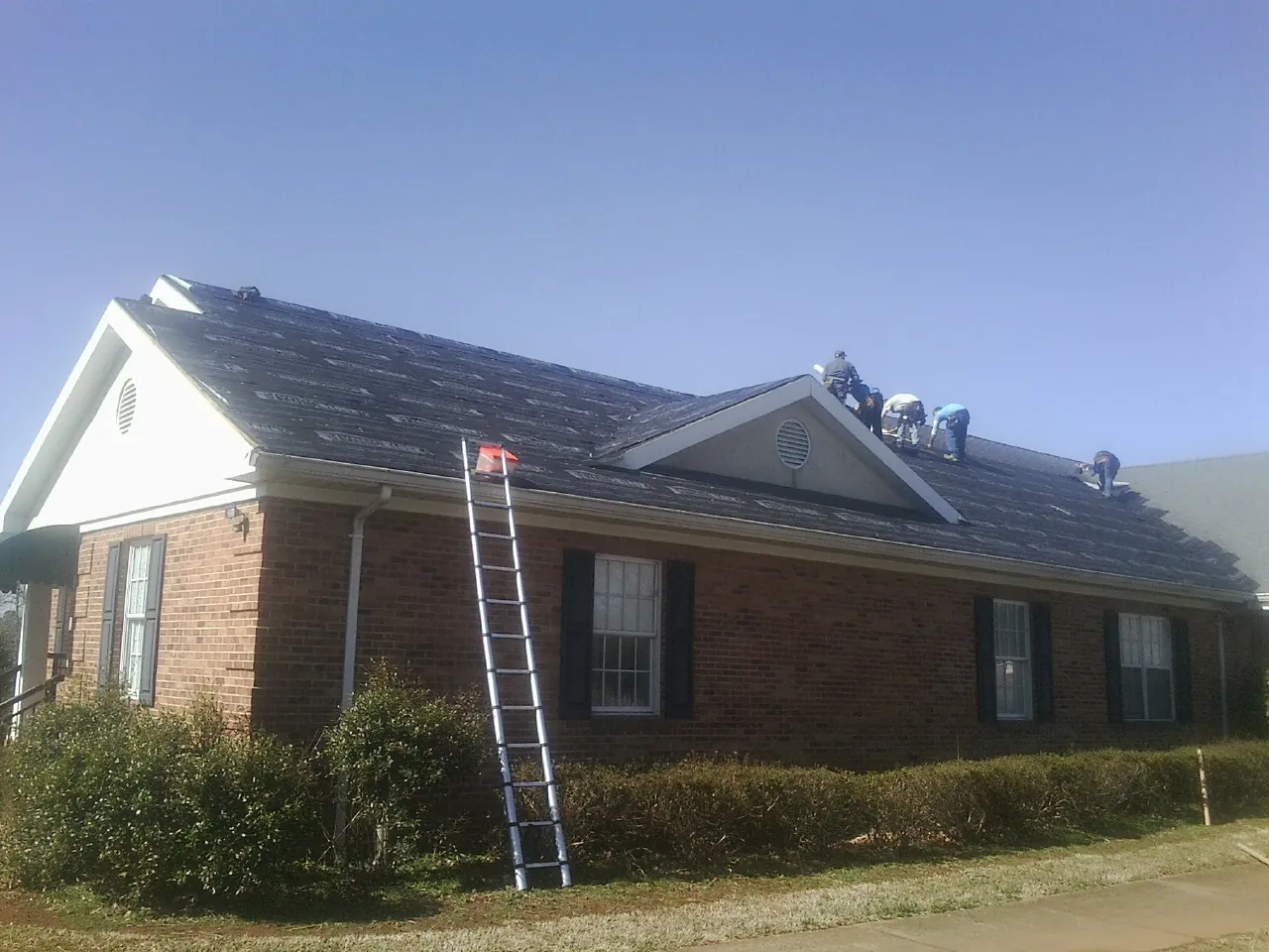 Crew tearing shingles off a brick ranch roof with a ladder set — Metal Roof Repair work by our crew in Jerico Springs
