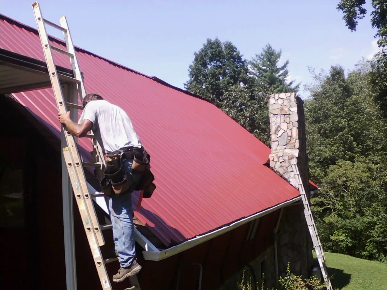 Roofer on a ladder beside a red metal roof and stone chimney — Soffit & Fascia Repair by Roofing Brothers in Jerico Springs